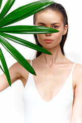 Fototapeta premium Elegant young woman obscured by vibrant green leaf, showcasing serene expression and minimalist style in bright setting. Perfect for lifestyle and beauty themes.
