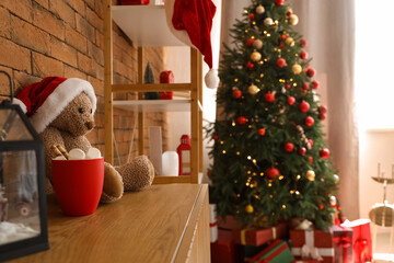 Obraz premium Toy bear with Santa hat and cup of cocoa on drawers in living room decorated for Christmas, closeup