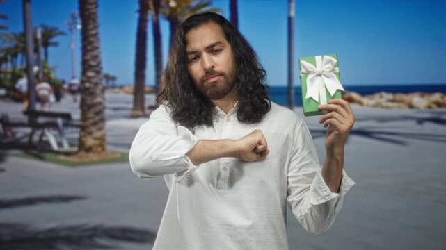 Man with long hair and beard holds green gift box aloft with right hand on a seaside street promenade lined with palm trees and benches; contemplation.