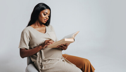 A beautiful pregnant woman is sitting comfortably on a chair and reading a book