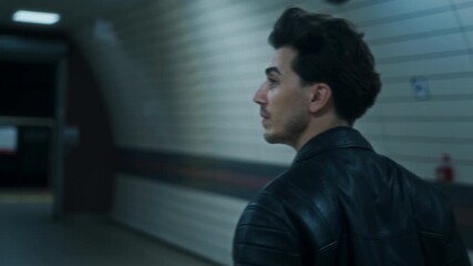 Handsome man in a leather jacket walks through a dimly lit underground subway tunnel, creating a cinematic urban night atmosphere.

