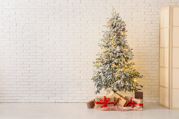 Decorated Christmas tree with glowing lights and gift boxes near white brick wall
