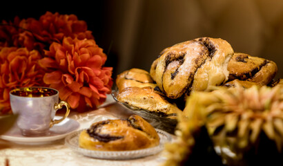Delicious homemade yeast dough pastries, among red flowers decorating the table, with a small cup. Beautiful and tasty. Served in warm colors. Sweet treats for the evening. Chocolate bun, without icin