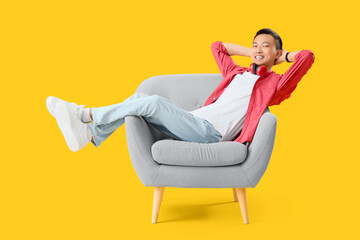 Fototapeta premium Young Asian man resting in grey armchair on yellow background