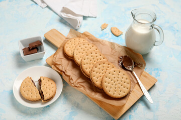 Board with tasty sandwich biscuits, chocolate cream and jug of milk on blue grunge background