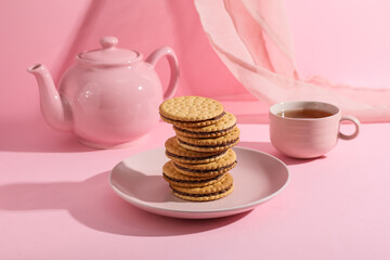 Plate with stack of tasty sandwich biscuits, teapot and cup of tea on pink background
