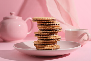 Plate with stack of tasty sandwich biscuits, teapot and cup of tea on pink background