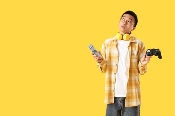 Thoughtful young Asian man with TV remote controller and gamepad on yellow background