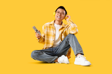 Fototapeta premium Young Asian man with TV remote controller and headphones sitting on yellow background