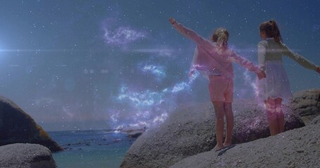 Fototapeta premium Standing sisters holding hands amid purple nebula glowing sparks on coastal boulders, copy space