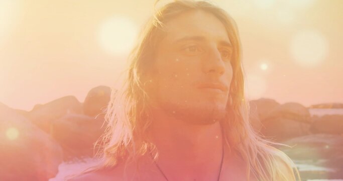 Gazing shirtless blonde-haired man standing amid rocky outcrops by sea at sunset, showing pendant - Powered by Adobe