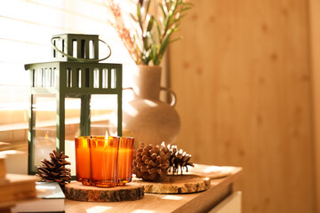 Burning candle, lantern and fir cones on chest of drawers in living room, closeup