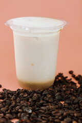 Close-up of an iced coffee or milk tea in a clear plastic cup on a bed of dark roasted coffee beans, set against a smooth coral studio background.