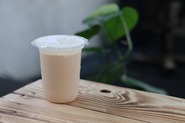 Sweating plastic cup of iced milk tea or coffee placed on a rustic wooden table, with water droplets, against a blurry green and gray outdoor background.