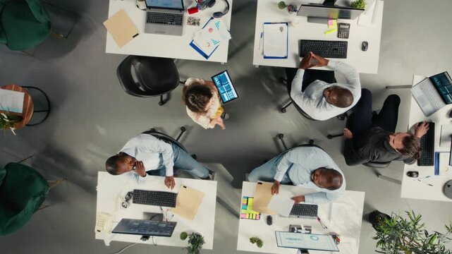 Top down view zoom in of colleagues in multinational company office discuss strategy, charts and analytics. Work team coordinates progress, insights and documentation for better outcomes. Camera A.