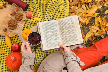 Young woman with cup of tea and cookies reading book on plaid in autumn park