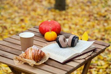 Book with headphones, croissant and cup of coffee on table in autumn park