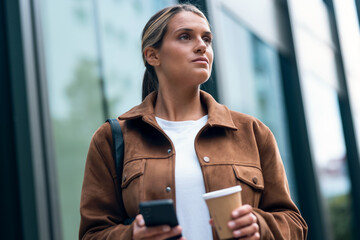 Pretty woman using her smartphone while drinking coffee walking through the city