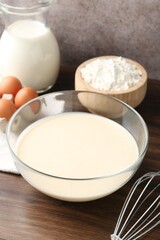 Liquid dough in bowl, whisk and ingredients on wooden table, closeup