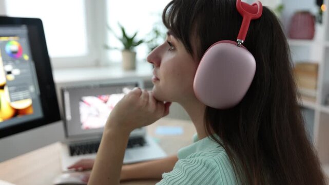 Professional retoucher putting on headphones and editing photo on computer at table in office. Camera moving