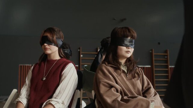 Group of blindfolded diverse teenagers sitting on chairs in circle and listening to psychotherapist speaking to them during hypnosis therapy session in sports hall