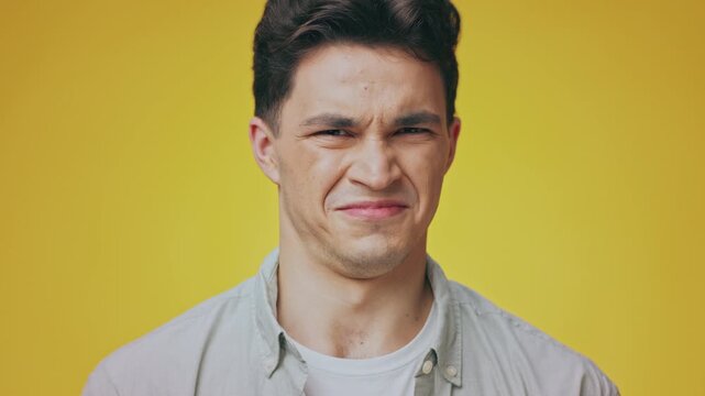 A young Caucasian man showcases expression with a scrunched face, showing disgust. The bright yellow backdrop enhances his charm and youthful spirit.