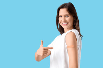 Fototapeta premium Happy young woman pointing at patch after vaccination on blue background. Immunity concept