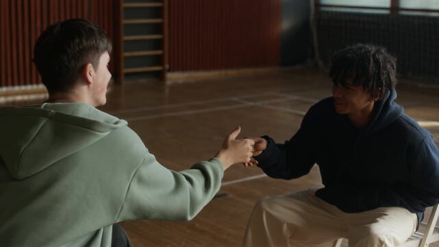 Two cheerful diverse teenage guys shaking hands during psychological session while sitting on chairs in sports hall and congratulating one another on successful therapy