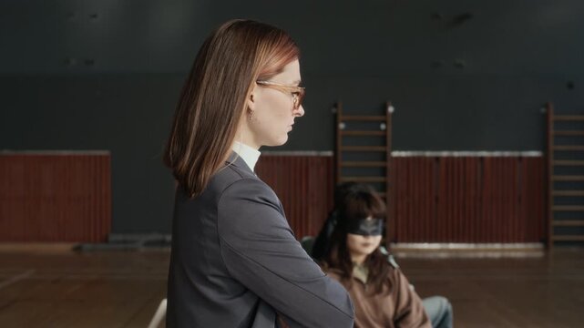 Young female psychotherapist with her arms crossed by chest walking along group of intercultural blindfolded teenage students sitting in circle and listening to her during hypnosis therapy