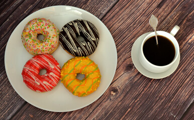 Four multi-colored glazed doughnuts with toppings on a round plate and a cup of hot black coffee on a wooden table.