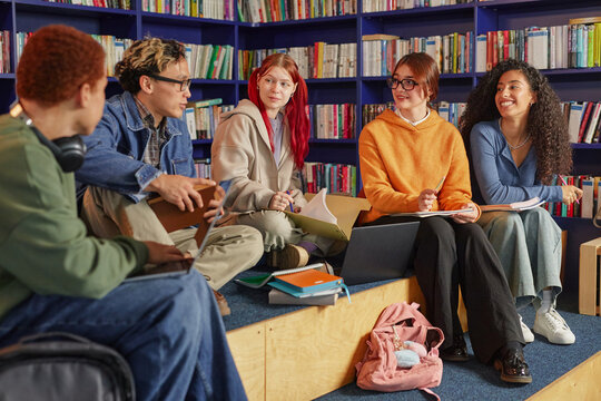 Group of diverse teenagers sitting together in library discussing schoolwork, multiethnic boys and girls interacting with notebooks and laptops, bookshelves in background