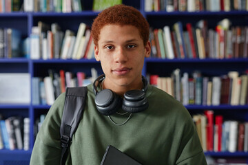Portrait of Biracial teenager boy standing in library holding laptop and wearing headphones around neck, looking directly at camera with neutral expression, bookshelves in background