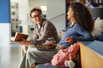 Asian young adult man smiling and holding book while talking to biracial young adult woman sitting beside him, both engaging in conversation with laptop and backpack visible