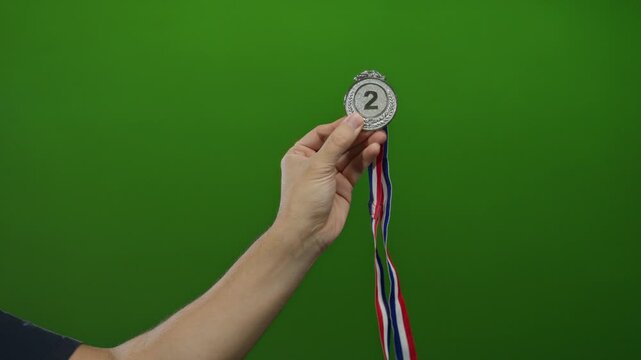 Male hand holding a silver medal with a colorful ribbon against a vibrant green background in an isolated setting, symbolizing achievement and success in a competitive environment.