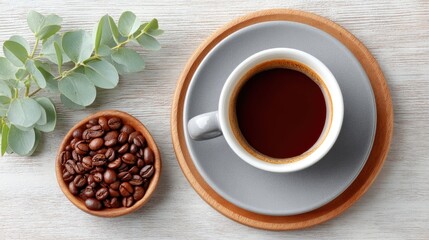 Top View Flat Lay of Roasted Coffee Beans in Wooden Bowl Next to Grey Cup of Black Coffee on White Wooden Table with Eucalyptus Sprig