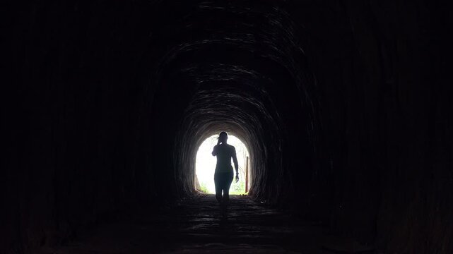 Woman Walks from Bright Tunnel Exit Toward the Camera into Deep Darkness in Full Silhouette Surrounded by Rough Rock Walls in a Symbolic Wide Perspective