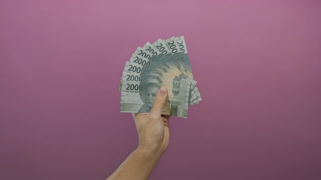 Man holding indonesian rupiah banknotes against a pink wall background, illustrating finance and wealth concept.