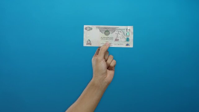 Male hand holds united arab emirates dirham banknote against a vibrant blue wall background, highlighting currency, finance, and economic concepts in an isolated setting.