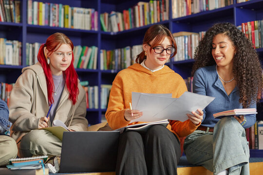 Three young adult women, including Caucasian and biracial individuals, sitting together in library holding papers and notebooks, collaborating on academic project, smiling and studying