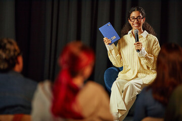 Young adult woman sitting on stage holding microphone and blue notebook, smiling and presenting to diverse audience, engaging in public speaking or workshop setting