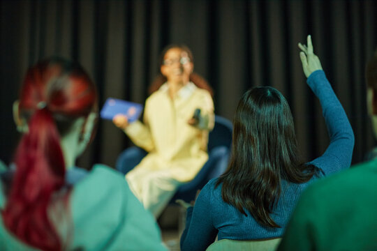 Teenage girl with brown hair sitting on stage holding tablet and remote, speaking to audience while teenage girl with dark hair raising hand in foreground, classroom or seminar setting - Powered by Adobe