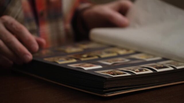 close-up philatelist hands carefully turning pages large album, looking at valuable collection postage stamps organized in protective sleeves on wooden table.