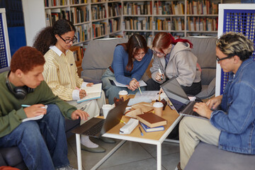 Group of multiethnic teenagers collaborating on project, using laptops and writing in notebooks...