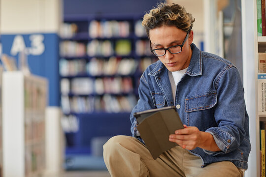 Young adult man sitting in library reading digital tablet, wearing glasses and focusing on screen, surrounded by bookshelves and blurred background - Powered by Adobe