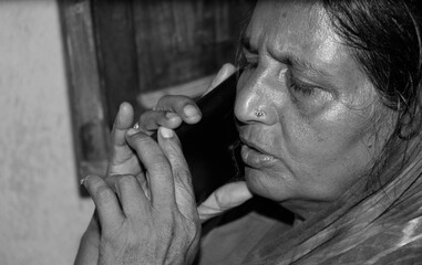 Black and white duotone photo shows a senior Indian pensioner at home, talking on her phone unaware...