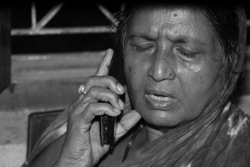 Monochrome image of a senior Indian woman talking on the phone, black and white background reflects vulnerability and loneliness as AI scammer targets the elderly in bank fraud.