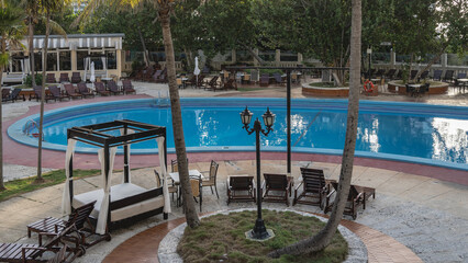 The resort swimming pool has a rounded curved shape.  Stairs with metal handrails, reflections on the blue water. Deck chairs, sun beds with mattresses and curtains in the poolside area.