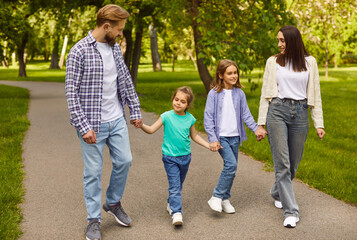 Happy family walking in summer park, parents and kids walk together along road, holding hands. Father and mother, son and daughter enjoying active stroll and talking with smile. Family time concept