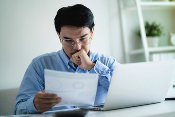 A man is sitting at a desk with a laptop and a stack of papers. He is looking at a piece of paper and he is in a state of deep thought