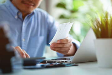 A man is sitting at a desk with a laptop and a stack of receipts. He is holding a receipt in his hand and he is organizing his finances